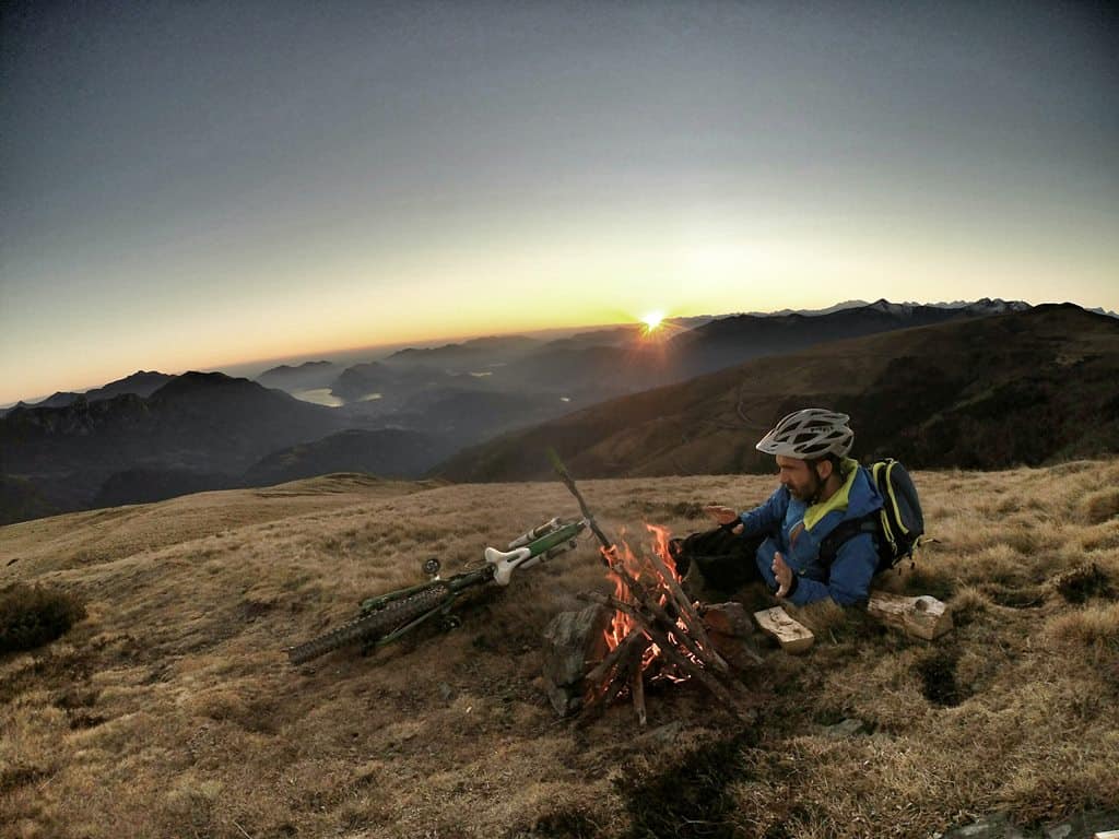 A man sitting next to a fire on top of a grass covered hillside with his trusty bike accessories.