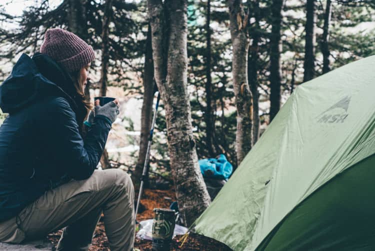 a person sitting next to a tent in the woods with a travel mug.