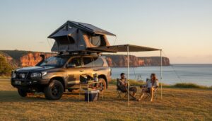Two people sit in camping chairs under an awning attached to an SUV with a rooftop tent, overlooking the ocean and cliffs at sunset. Camping gear is set up beside them on grassy terrain.