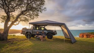 Two people sit under an awning attached to a parked SUV on a grassy cliff overlooking the ocean at sunset, with dramatic clouds and a large tree nearby.