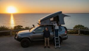 A man stands beside an SUV parked by the ocean at sunset, while a woman climbs a ladder to a rooftop tent on the vehicle. The sun is low over the calm water, casting a warm glow.