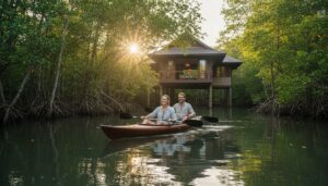 A smiling couple paddles a kayak through calm water surrounded by lush mangroves, with a wooden house on stilts in the background and sunlight streaming through the trees.
