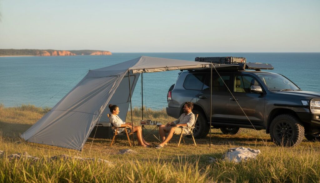 Two people sit in camping chairs under an awning attached to a parked SUV, overlooking the ocean and cliffs in the distance on a sunny day. Camping gear is visible on the roof of the vehicle.